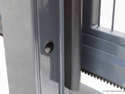 Billedet viser et nærbillede af en grå Skydeport "V-Star EXTREME" 2D Panelfyld 8/6/8 MM automatisk skydeportsystem, med en lodret stolpe med en rund sensor eller bolt og vandrette stænger på porten. En gearstang er delvist synlig i bunden af porten.
