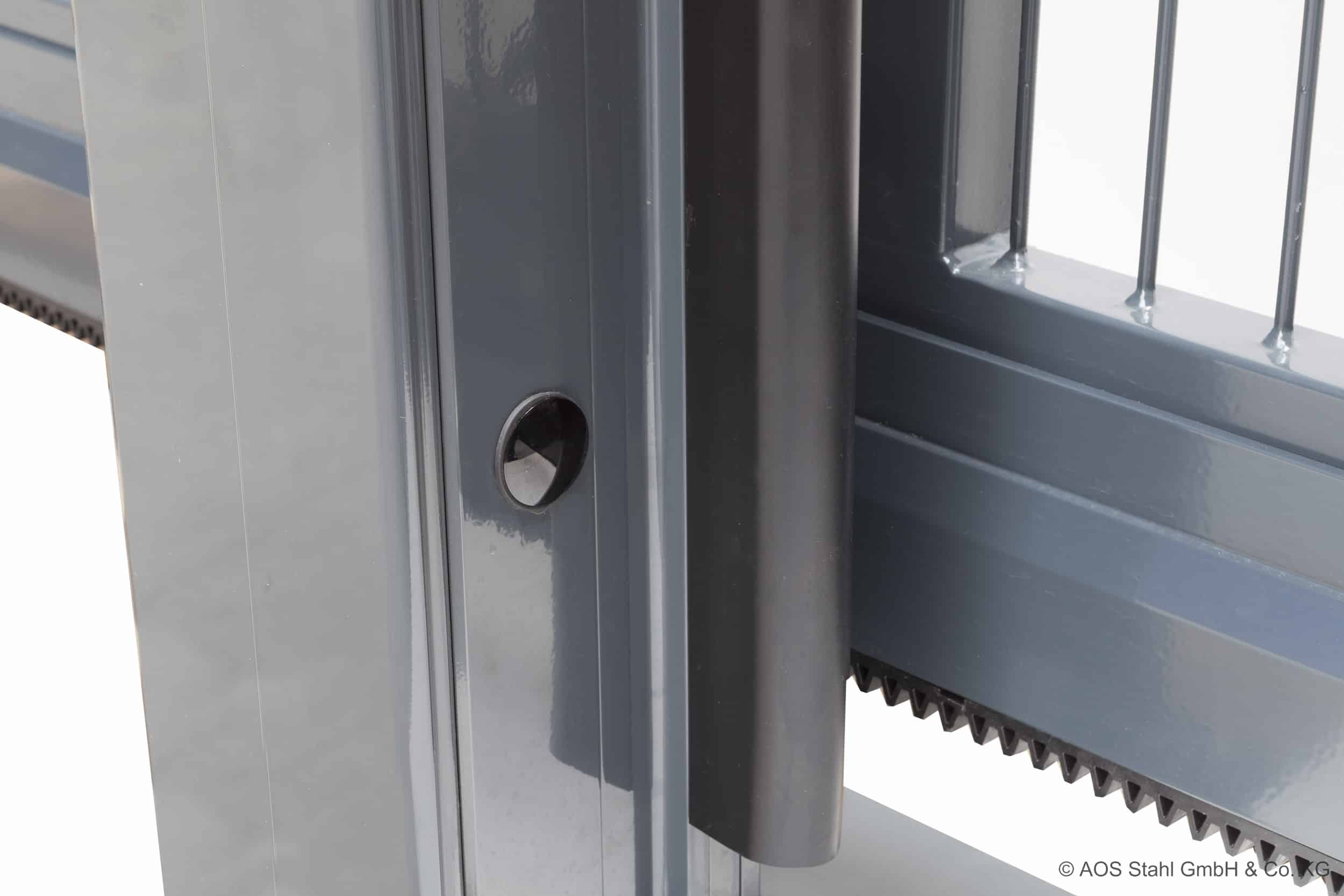 Billedet viser et nærbillede af en grå Skydeport "V-Star EXTREME" 2D Panelfyld 8/6/8 MM automatisk skydeportsystem, med en lodret stolpe med en rund sensor eller bolt og vandrette stænger på porten. En gearstang er delvist synlig i bunden af porten.