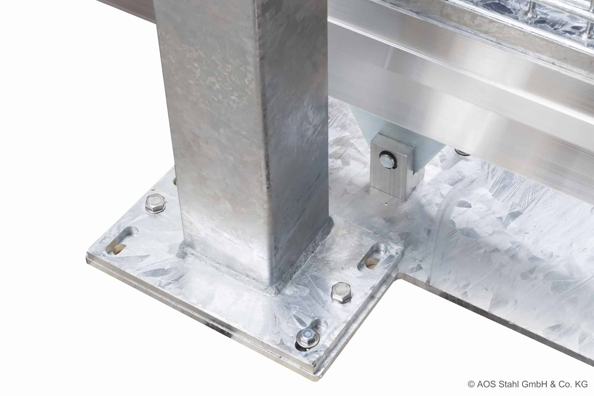 Nærbillede af en metallisk struktur med en lodret stålstolpe boltet til en flad bundplade, der ligner det robuste design, der findes i Skydeport "V-Star EXTREME" 2D Panelfyld 8/6/8 MM produkter. Bundpladen er fastgjort med fire bolte, og metaloverfladen har en galvaniseret finish. Baggrunden indeholder yderligere metalliske komponenter © AOS Stahl GmbH & Co. KG.