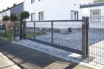 Et moderne bolighus med en slank Skydeport "V-Star EXTREME" 2D Panelfyld 8/6/8 MM metalport og hegn. Indkørslen er brolagt med grå mursten, der strækker sig til en lille have prydet med frodigt grønt. Husets ydre er hvidt med store vinduer, hvilket giver et rent og moderne udseende.