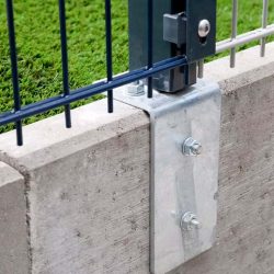 Nærbillede af en Stålstålper 60x40mm med dækliste TYPE KA Komplet metalhegnplade monteret på betonoverflade ved hjælp af metalbeslag med to bolte. Grønne planter kan ses i baggrunden, delvist synlige gennem hegnet. Konstruktionen fremstår robust med sikker forbindelse mellem hegnet og beton.