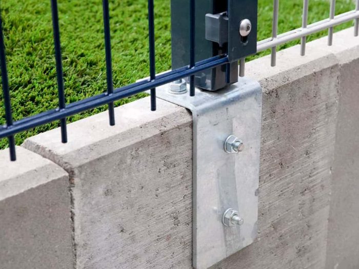 Nærbillede af en Stålstålper 60x40mm med dækliste TYPE KA Komplet metalhegnplade monteret på betonoverflade ved hjælp af metalbeslag med to bolte. Grønne planter kan ses i baggrunden, delvist synlige gennem hegnet. Konstruktionen fremstår robust med sikker forbindelse mellem hegnet og beton.