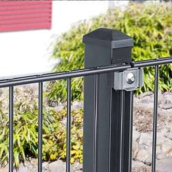 Et sort metalhegn med lodrette stænger er fastgjort til en robust sort stolpe ved hjælp af et sølvbeslag, toppet med Stolpehat 60x40mm Plast TYPE U. Baggrunden har grønne buske og slørede elementer, inklusive en rød vinduesskodder. Sten er synlig ved bunden af hegnet.