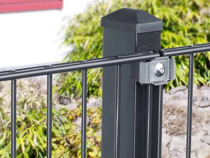 Et sort metalhegn med lodrette stænger er fastgjort til en robust sort stolpe ved hjælp af et sølvbeslag, toppet med Stolpehat 60x40mm Plast TYPE U. Baggrunden har grønne buske og slørede elementer, inklusive en rød vinduesskodder. Sten er synlig ved bunden af hegnet.