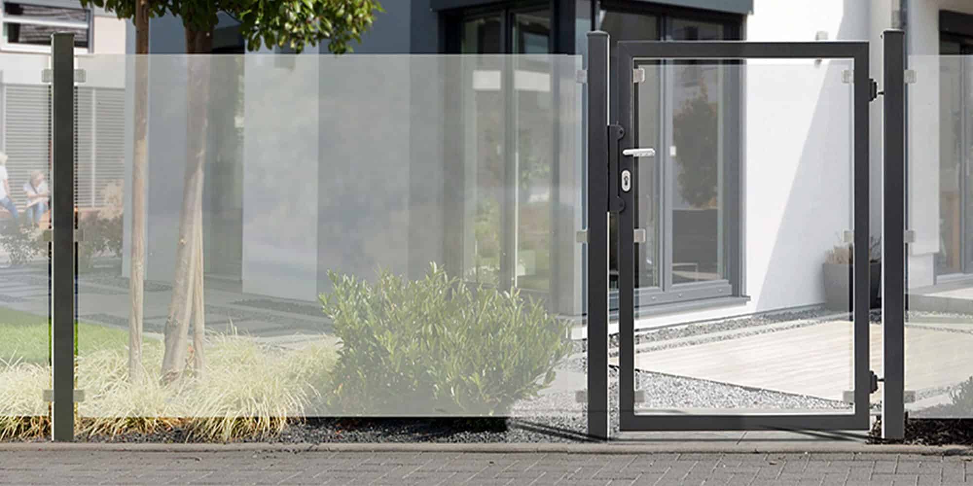 En moderne indgang til huset har et minimalistisk design med et hegn og låge af klart glas, indrammet i slankt gråt metal. Hegnet grænser op til en lille, velholdt græsplæne med en grøn busk, der fører til en brolagt gangbro og husets store vinduespartier, hvilket sikrer nem vedligeholdelse af hegn.