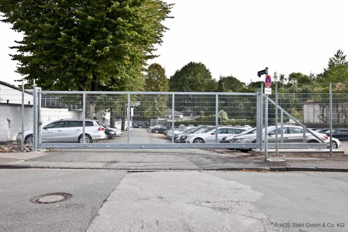 En stor og sikker Skydeport "EasyStar" port, bestående af Lodret Gitter paneler, omslutter en parkeringsplads fyldt med biler. Metalhegnet spænder over hele indgangsbredden på 3 til 9,5 meter. Træer og bygninger er synlige i baggrunden, med parkeringsregler på Skydeport-panelerne.