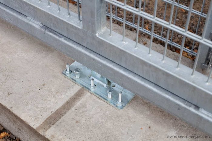 Et nærbillede af en metalhegnspæls base fastgjort til et betonfundament med flere bolte. Metalpladen har fire bolte stramt skruet ind i, hvilket sikrer stabiliteten af Skydeport “EasyStar” – Lodret Gitter eller Panel hegns fyld. Jorden er dækket af små grus.