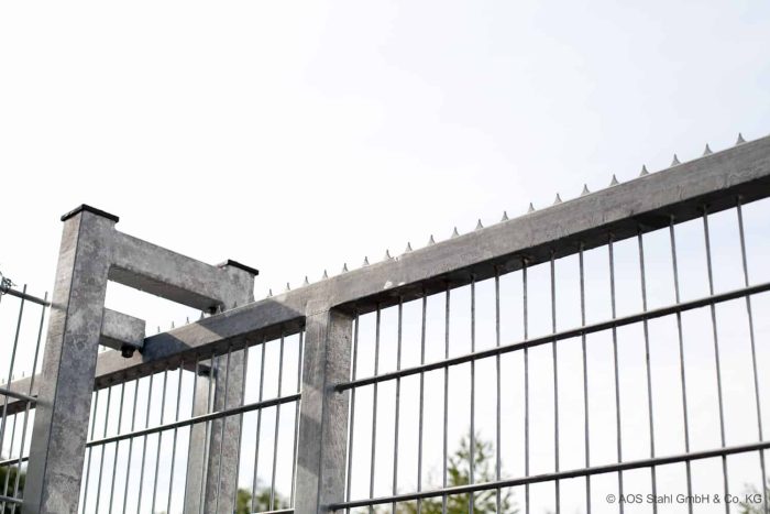 Et nærbillede af et højt Skydeport "EasyStar" metalhegn med pigge under en klar himmel. Hegnet viser lodrette stænger og skarpe pigge på toppen for øget sikkerhed. Sløret grønt er synligt i baggrunden. Et vandmærke i nederste højre hjørne lyder "© AOS Stahl GmbH & Co. KG.