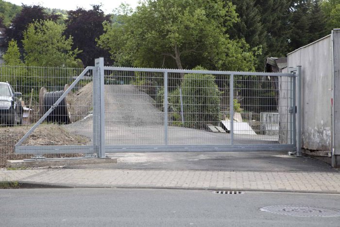 En stor, lukket Skydeport "EasyStar" port med hegns fyld design spærrer indgangen til en grus indkørsel omgivet af grønne områder. Til venstre er en del af et parkeret køretøj synligt. Porten er fastgjort til en betonmur til højre, og scenen er sat udendørs på en solskinsdag.