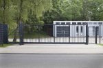 Ved indgangen til en asfalteret indkørsel står en Skydeport "EasyStar"-port, med lodret gitter eller panelhegn, i slankt sort metal. Bag den lukkede port ligger en moderne etplansbygning med store vinduespartier. Bygningen er omgivet af frodige grønne træer og græs. Vejen i forgrunden er tom.