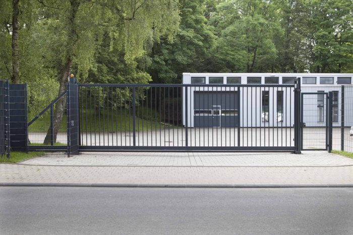 Ved indgangen til en asfalteret indkørsel står en Skydeport "EasyStar"-port, med lodret gitter eller panelhegn, i slankt sort metal. Bag den lukkede port ligger en moderne etplansbygning med store vinduespartier. Bygningen er omgivet af frodige grønne træer og græs. Vejen i forgrunden er tom.