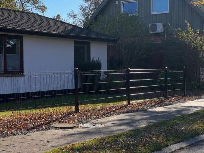 Et sort metalhegn adskiller et hvidt hus og en grøn græsplæne fra et fortov dækket af nedfaldne blade. Et andet hus og træer er synlige i baggrunden under en klar himmel.