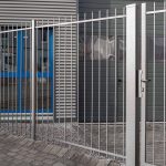 Baluster Hegn Pulverlakeret Antracit Metal – komplet med stålstolper og beslag – står sikkert låst foran en moderne bygning, der kan prale af en grå facade og vinduer indrammet i blåt. Den murstensbelagte grund komplementerer bygningens entré, som er flankeret af slanke balustre ved siden af vinduerne.