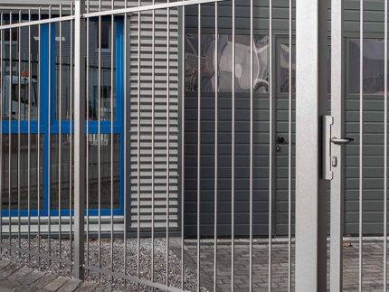 Baluster Hegn Pulverlakeret Antracit Metal – komplet med stålstolper og beslag – står sikkert låst foran en moderne bygning, der kan prale af en grå facade og vinduer indrammet i blåt. Den murstensbelagte grund komplementerer bygningens entré, som er flankeret af slanke balustre ved siden af vinduerne.