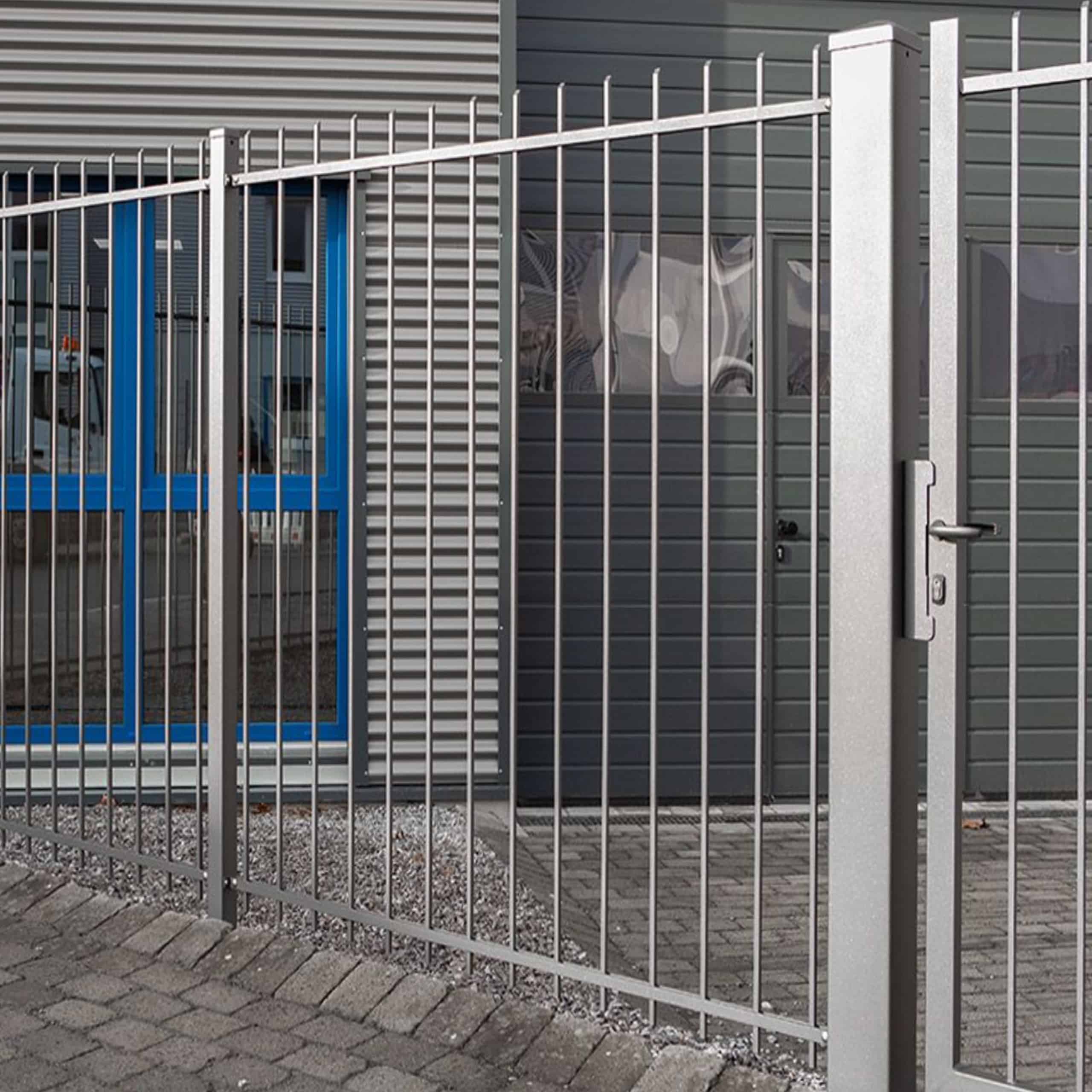 Baluster Hegn Pulverlakeret Antracit Metal – komplet med stålstolper og beslag – står sikkert låst foran en moderne bygning, der kan prale af en grå facade og vinduer indrammet i blåt. Den murstensbelagte grund komplementerer bygningens entré, som er flankeret af slanke balustre ved siden af vinduerne.