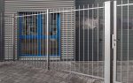 En Baluster Hegn Pulverlakeret Antracit Metal-port, komplet med stålstolper og beslag, er lukket foran en bygningsindgang. Bygningen har et gråt metal udvendigt med to blå trimvinduer og et bølgeformet design. Jorden er brolagt med gråsten, der hænger sammen, og der er grus i nærheden af porten.