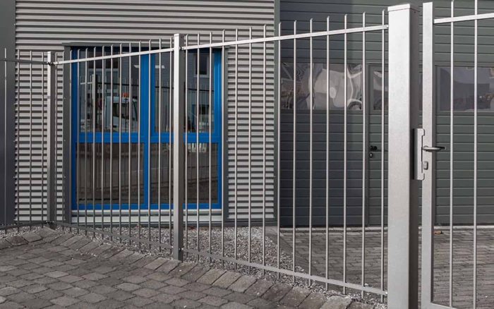 En Baluster Hegn Pulverlakeret Antracit Metal-port, komplet med stålstolper og beslag, er lukket foran en bygningsindgang. Bygningen har et gråt metal udvendigt med to blå trimvinduer og et bølgeformet design. Jorden er brolagt med gråsten, der hænger sammen, og der er grus i nærheden af porten.