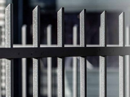 Nærbillede af Baluster Hegn Pulverlakeret Antracit Metal - Inkl. Stålstolper & Beslag, med lodrette stænger. De mørke metalstænger er jævnt fordelt og har en let struktureret overflade. Baggrunden er sløret, hvilket fremhæver hegnets struktur.