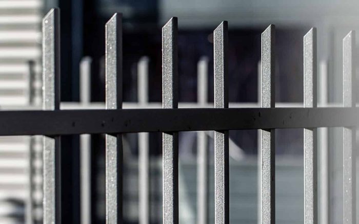 Nærbillede af Baluster Hegn Pulverlakeret Antracit Metal - Inkl. Stålstolper & Beslag, med lodrette stænger. De mørke metalstænger er jævnt fordelt og har en let struktureret overflade. Baggrunden er sløret, hvilket fremhæver hegnets struktur.