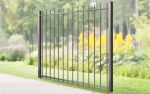 Baluster Hegn Pulverlakeret Antracit Metal, komplet med stålstolper og beslag, er installeret i et frodigt, grønt udendørsområde omgivet af træer og forskellig vegetation. Denne elegante port strækker sig over en gangbro og skaber en barriere i det naturlige landskab. Lyse gule blomster er synlige i baggrunden.