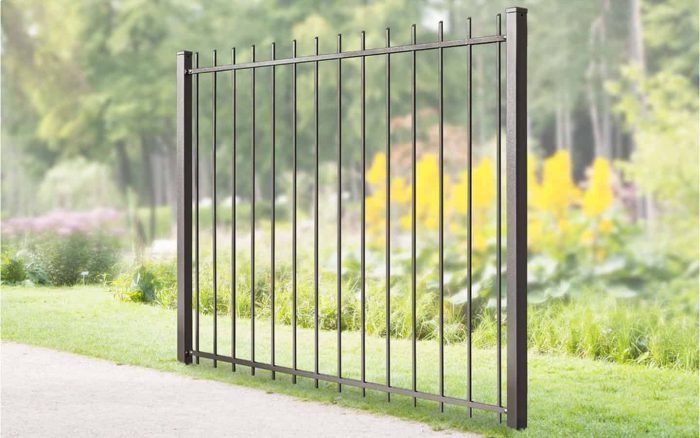 Baluster Hegn Pulverlakeret Antracit Metal, komplet med stålstolper og beslag, er installeret i et frodigt, grønt udendørsområde omgivet af træer og forskellig vegetation. Denne elegante port strækker sig over en gangbro og skaber en barriere i det naturlige landskab. Lyse gule blomster er synlige i baggrunden.