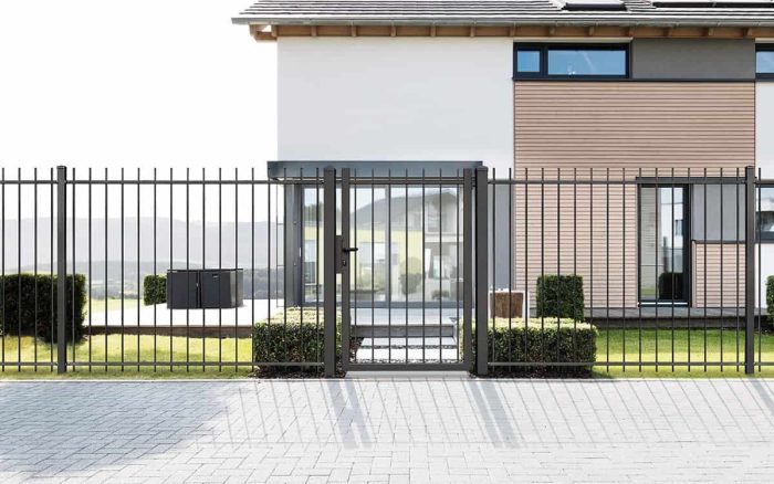 Et moderne hus med en kombination af hvid- og træpaneler kan prale af store vinduer og frodige grønne plæner. På tværs af fronten er der et Baluster Hegn Pulverlakeret Antracit Metal hegn komplet med lodrette stænger, stålstolper og beslag, der giver en slank grænse. Firkantede buske flankerer begge sider af porten for ekstra elegance.
