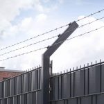 Et nærbillede af et højt, sort metalsikringshegn toppet med vinklede sektioner og linjer af pigtråd holdt af Pigtrådsholder Enkelt 45° - 3 tråde til 60x40mm stolpe. I baggrunden er en overskyet himmel delvist synlig.