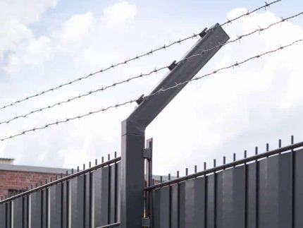 Et nærbillede af et højt, sort metalsikringshegn toppet med vinklede sektioner og linjer af pigtråd holdt af Pigtrådsholder Enkelt 45° - 3 tråde til 60x40mm stolpe. I baggrunden er en overskyet himmel delvist synlig.