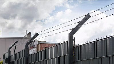 På baggrund af en overskyet himmel fremviser et højt sort metalhegn toppet med pigtråd den robuste Pigtrådsholder Enkelt 45° - 3 tråde til 60x40mm stolpe. De kantede metalstolper, der understøtter wiren, giver et delvist overblik over nogle industribygninger bag hegnet.