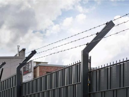 På baggrund af en overskyet himmel fremviser et højt sort metalhegn toppet med pigtråd den robuste Pigtrådsholder Enkelt 45° - 3 tråde til 60x40mm stolpe. De kantede metalstolper, der understøtter wiren, giver et delvist overblik over nogle industribygninger bag hegnet.