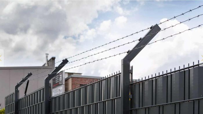 På baggrund af en overskyet himmel fremviser et højt sort metalhegn toppet med pigtråd den robuste Pigtrådsholder Enkelt 45° - 3 tråde til 60x40mm stolpe. De kantede metalstolper, der understøtter wiren, giver et delvist overblik over nogle industribygninger bag hegnet.