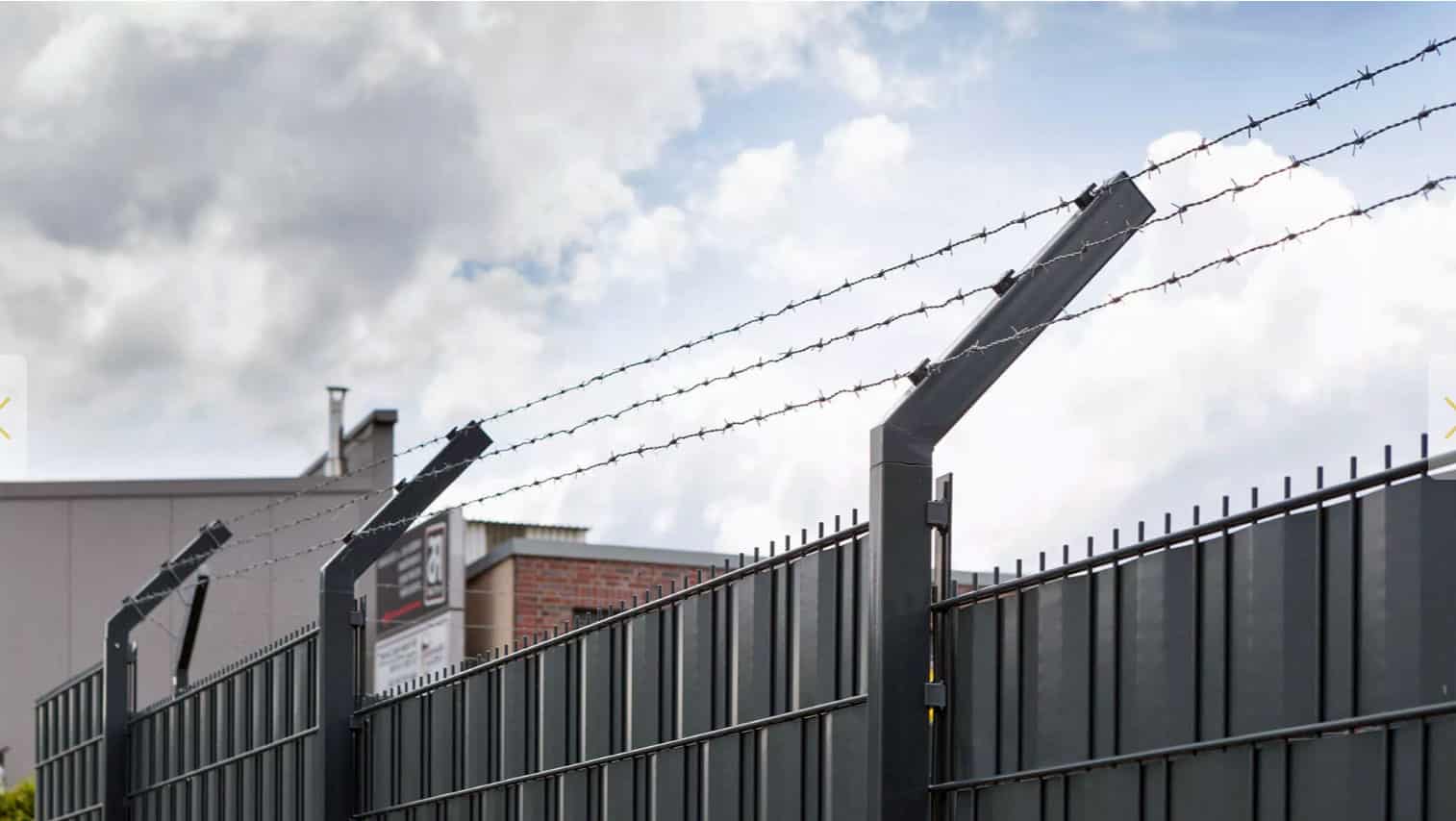 På baggrund af en overskyet himmel fremviser et højt sort metalhegn toppet med pigtråd den robuste Pigtrådsholder Enkelt 45° - 3 tråde til 60x40mm stolpe. De kantede metalstolper, der understøtter wiren, giver et delvist overblik over nogle industribygninger bag hegnet.