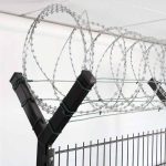 Nærbillede af et sort sikkerhedshegn toppet med spoler af barbertråd. Barberwiren, der holdes sikkert af Pigtrådsholder Dobbelt 45° - 6 tråde til 60x40mm stolpe, er arrangeret i jævnt fordelte løkker langs toppen af hegnet, hvilket sandsynligvis tjener som en afskrækkelse mod klatring eller uautoriseret adgang. Baggrunden viser et hvidt loft.