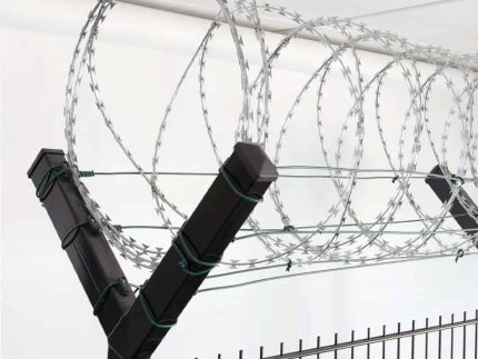 Nærbillede af et sort sikkerhedshegn toppet med spoler af barbertråd. Barberwiren, der holdes sikkert af Pigtrådsholder Dobbelt 45° - 6 tråde til 60x40mm stolpe, er arrangeret i jævnt fordelte løkker langs toppen af hegnet, hvilket sandsynligvis tjener som en afskrækkelse mod klatring eller uautoriseret adgang. Baggrunden viser et hvidt loft.