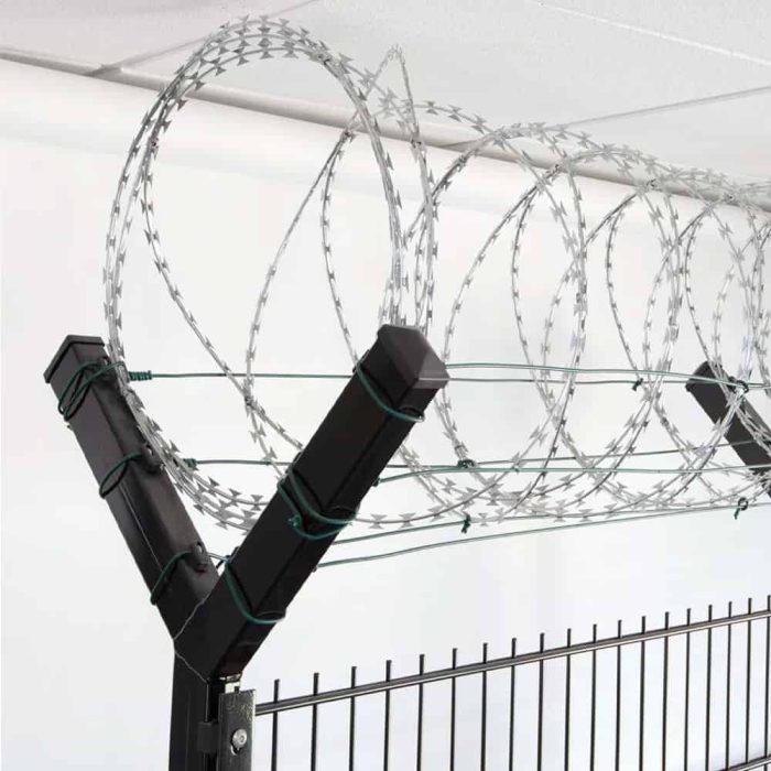 Nærbillede af et sort sikkerhedshegn toppet med spoler af barbertråd. Barberwiren, der holdes sikkert af Pigtrådsholder Dobbelt 45° - 6 tråde til 60x40mm stolpe, er arrangeret i jævnt fordelte løkker langs toppen af hegnet, hvilket sandsynligvis tjener som en afskrækkelse mod klatring eller uautoriseret adgang. Baggrunden viser et hvidt loft.