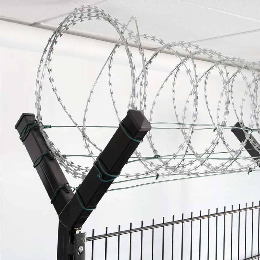 Nærbillede af et sort sikkerhedshegn toppet med spoler af barbertråd. Barberwiren, der holdes sikkert af Pigtrådsholder Dobbelt 45° - 6 tråde til 60x40mm stolpe, er arrangeret i jævnt fordelte løkker langs toppen af hegnet, hvilket sandsynligvis tjener som en afskrækkelse mod klatring eller uautoriseret adgang. Baggrunden viser et hvidt loft.