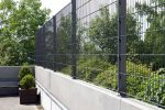 Et højt metalhegn med netdesign, pulverlakeret i RAL 9005, er installeret oven på en betonvæg. Hegnet omslutter et udendørs område med frodigt grønt og grønne træer i baggrunden. På venstre side af billedet er der en plantekasse med en lille busk ved siden af en 60x40mm Stolpe fastgjort ved hjælp af Stolpefod til 60x40mm Stolpe Sort RAL 9005 - 4 huller.