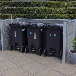 Tre sorte skraldespande med farvekodede klistermærker står på rad og række på en brolagt overflade, omsluttet af et træ- og metalhegn med stålstolper. Hække ses i baggrunden, og en potteplante er placeret til højre for skraldespandene.