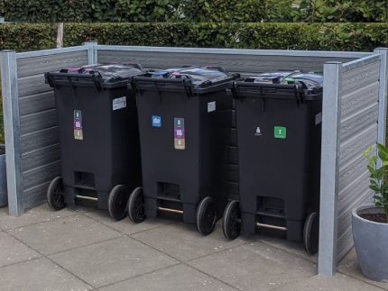 Tre sorte skraldespande med farvekodede klistermærker står på rad og række på en brolagt overflade, omsluttet af et træ- og metalhegn med stålstolper. Hække ses i baggrunden, og en potteplante er placeret til højre for skraldespandene.