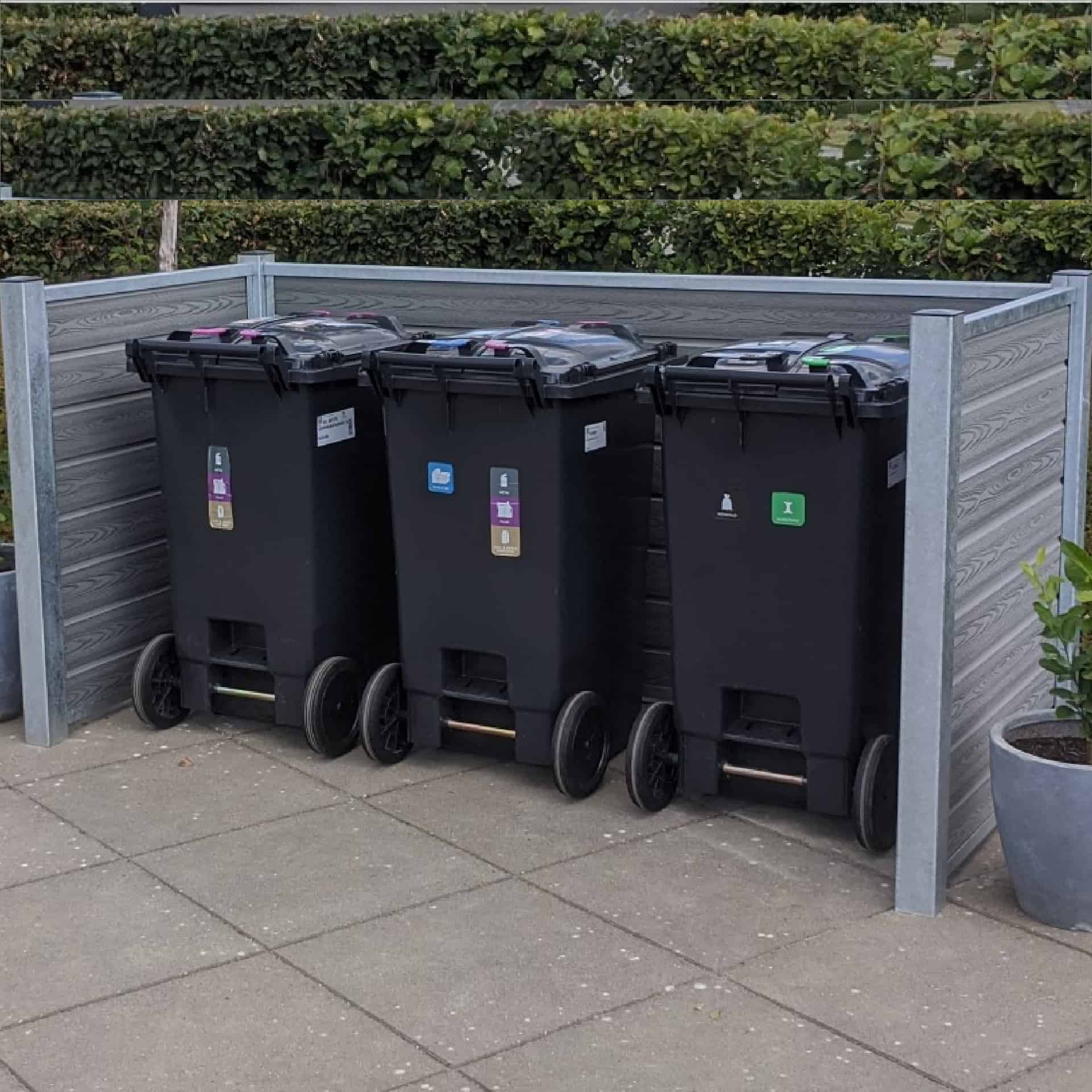 Tre sorte skraldespande med farvekodede klistermærker står på rad og række på en brolagt overflade, omsluttet af et træ- og metalhegn med stålstolper. Hække ses i baggrunden, og en potteplante er placeret til højre for skraldespandene.
