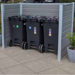 Tre sorte genbrugsspande med farverige klistermærker, der angiver forskellige typer affald, står opstillet ved siden af hinanden i en Grå komposit skraldespandsskjul understøttet af stålstolper. Indhegningen placeres på en flisebelagt overflade med potteplanter på hver side.