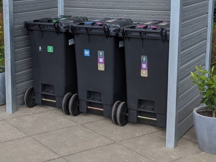 Tre sorte genbrugsspande med farverige klistermærker, der angiver forskellige typer affald, står opstillet ved siden af hinanden i en Grå komposit skraldespandsskjul understøttet af stålstolper. Indhegningen placeres på en flisebelagt overflade med potteplanter på hver side.