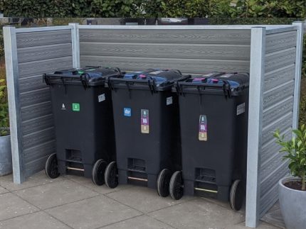 Tre sorte skraldespande på hjul står side om side inden for en grå komposit skraldespandsskjul på et brolagt område. Hver beholder har et farvet klistermærke på fronten til sortering af forskellige typer affald. Grønt og hække ses i baggrunden, indrammet af robuste stålstolper for ekstra støtte.