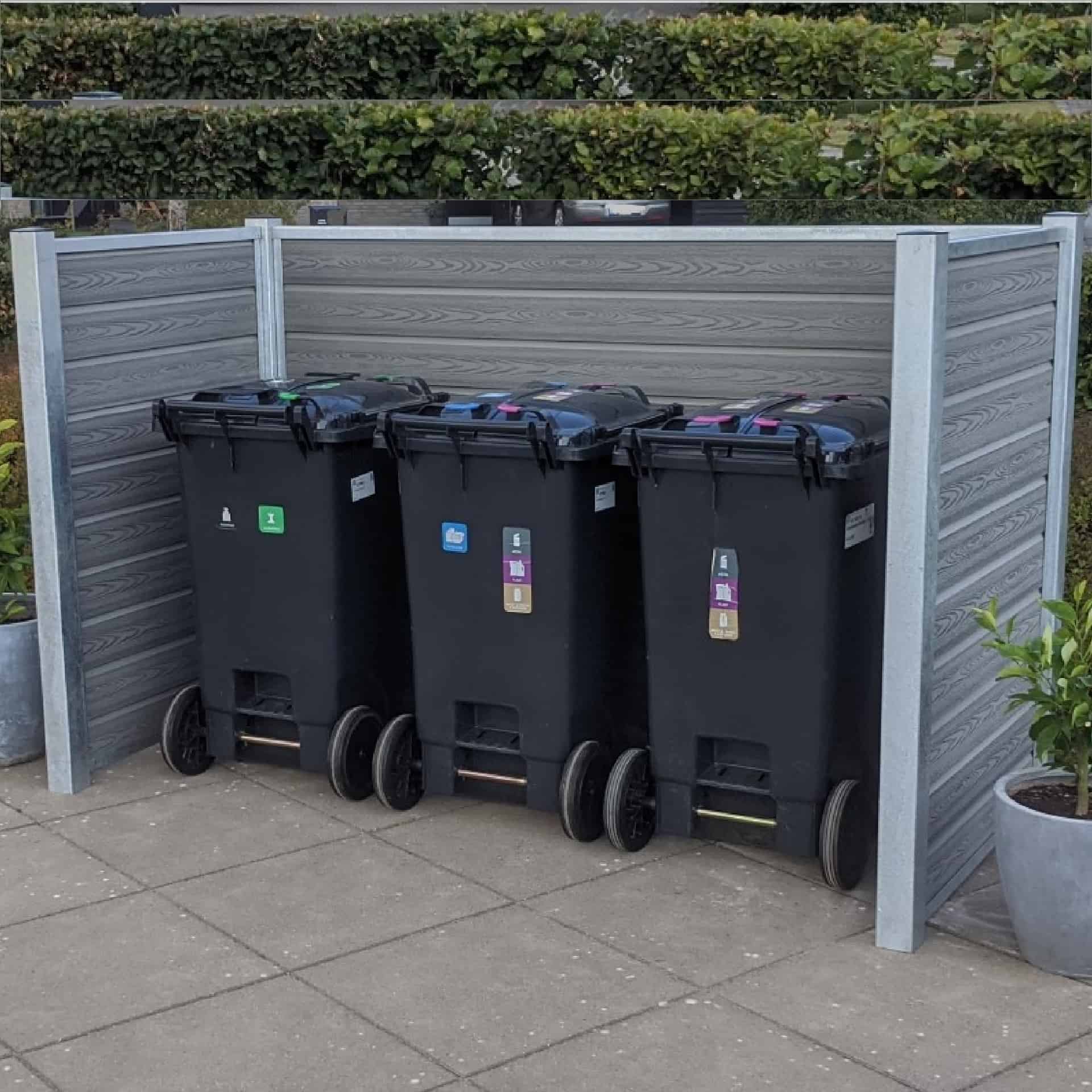 Tre sorte skraldespande på hjul står side om side inden for en grå komposit skraldespandsskjul på et brolagt område. Hver beholder har et farvet klistermærke på fronten til sortering af forskellige typer affald. Grønt og hække ses i baggrunden, indrammet af robuste stålstolper for ekstra støtte.
