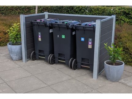 Tre sorte skraldespande med forskellige farvede låg er placeret side om side i en Komposit skraldespandsskjul understøttet af stålstolper. Området er flisebelagt med lys grå beton, og to store grå krukker med små grønne planter flankerer børstets sorteringsspande.
