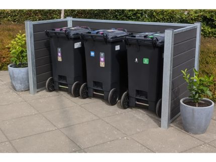 Tre sorte skraldespande på hjul med farvede låg er pænt arrangeret i en børstet sort Komposit skraldespandsskjul. To potteplanter sidder i hjørnerne af indhegningen, som er sat på en flisebelagt terrasse med grønt løv i baggrunden.