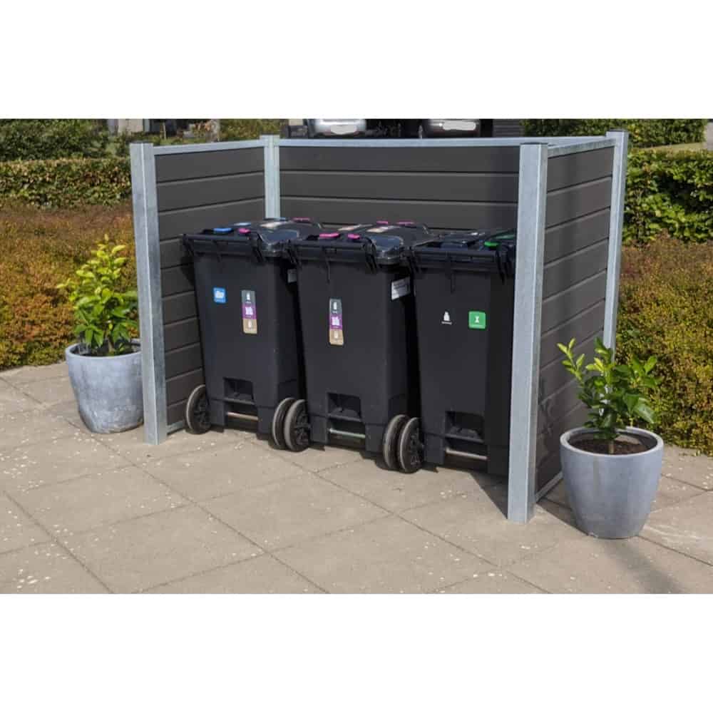 Et sæt med tre sorte skraldespande placeret i en skraldespandsskjul med stålstolper udendørs. Hver beholder har forskellige farvede låg og etiketter til genbrugskategorier. På hver side af børstet sorteringsspande står store potteplanter. Området er belagt med grå fliser og omgivet af grønt.