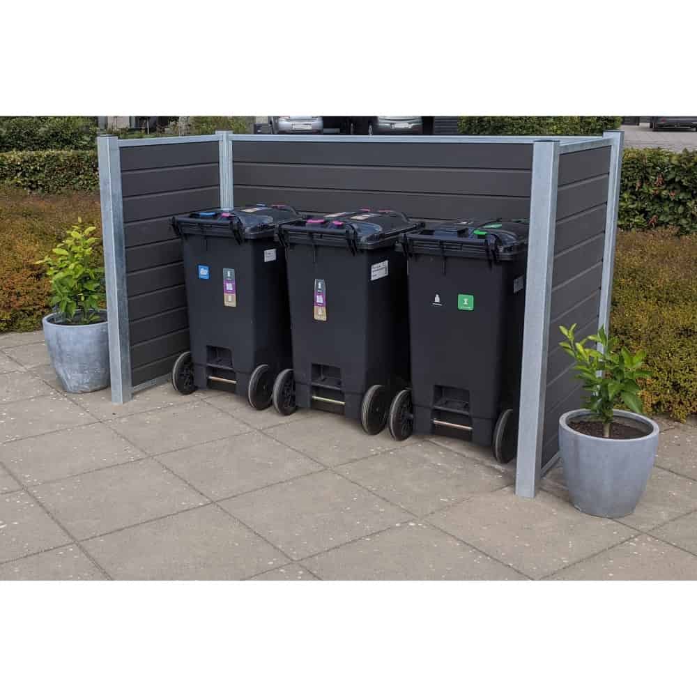 Tre sorte skraldespande er opstillet i en pæn Komposit skraldespandsskjul med grå panelvægge understøttet af stålstolper. Hver beholder har en forskellig farvet etiket, der angiver genbrug, almindeligt affald og organisk affald. To potteplanter er placeret på hver side af indhegningen, og buske er i baggrunden.
