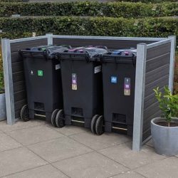 Et sæt med tre sorte skraldespandsspande, hver med et forskelligt farvet låg og etiket, er placeret i en pæn træ-skraldespandsskjul udendørs. Beholderne hviler på en flisebelagt overflade med potteplanter i nærheden og grønne hække i baggrunden, alt sammen understøttet af robuste stålstolper.