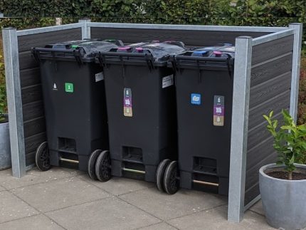 Et sæt med tre sorte skraldespandsspande, hver med et forskelligt farvet låg og etiket, er placeret i en pæn træ-skraldespandsskjul udendørs. Beholderne hviler på en flisebelagt overflade med potteplanter i nærheden og grønne hække i baggrunden, alt sammen understøttet af robuste stålstolper.
